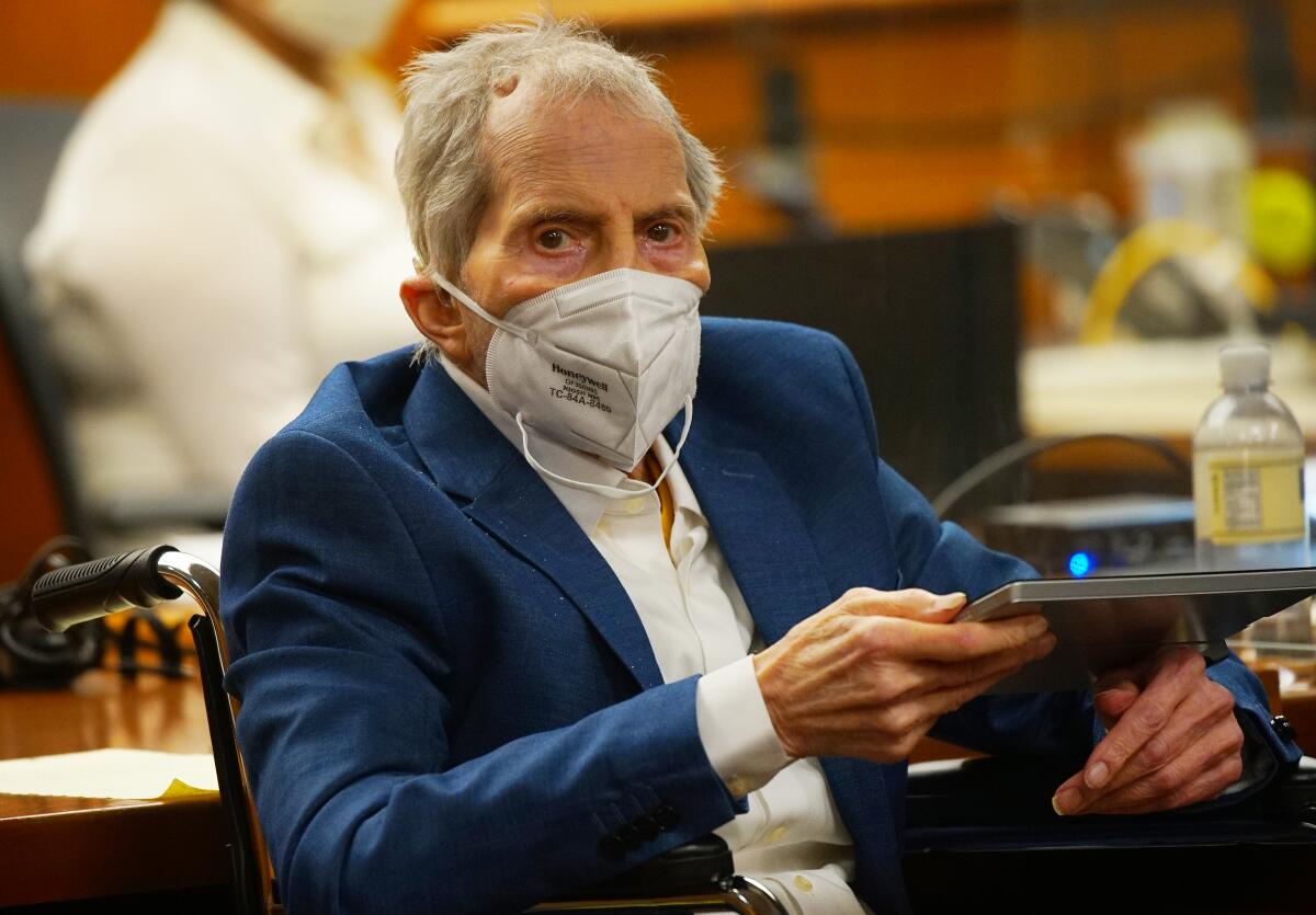 A gray-haired man in a blue suit jacket and wearing a face mask holds a tablet as he sits in a wheelchair