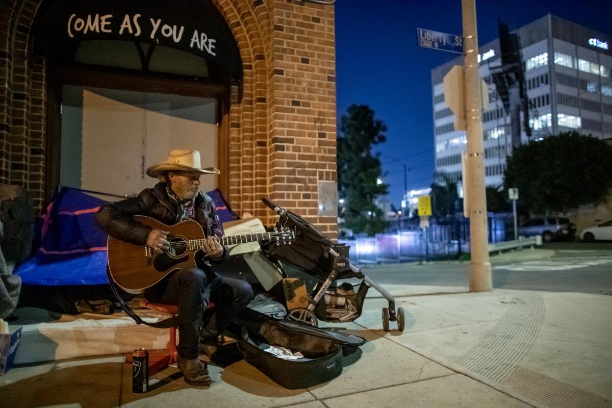 Man wearing a cowboy hat sits outside in a city playing the guitar