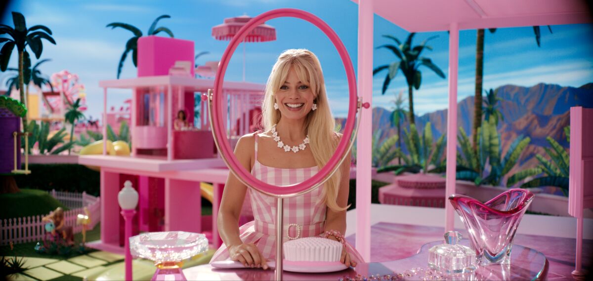 Margot Robbie as Barbie is smiling in front of a mirror inside a pink doll house