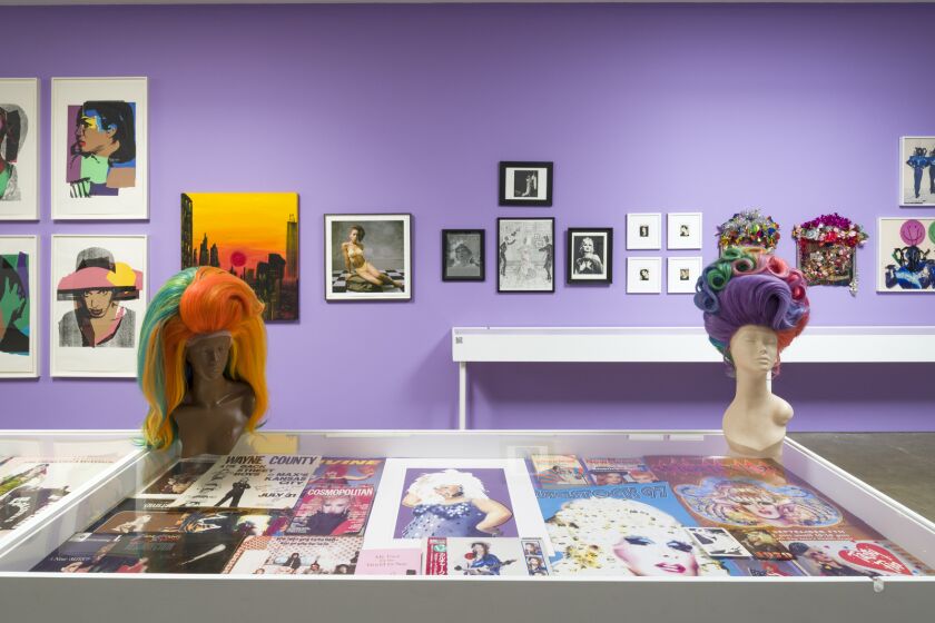 A gallery with lavender walls contains vitrines with drag ephemera and two mannequins wearing flamboyant rainbow wigs