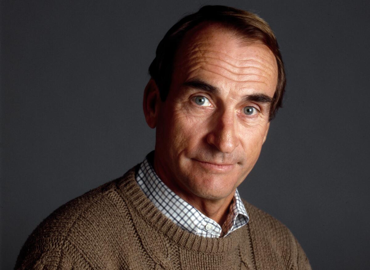 A man in a windowpane plaid shirt and brown pullover sweater looks at the camera.