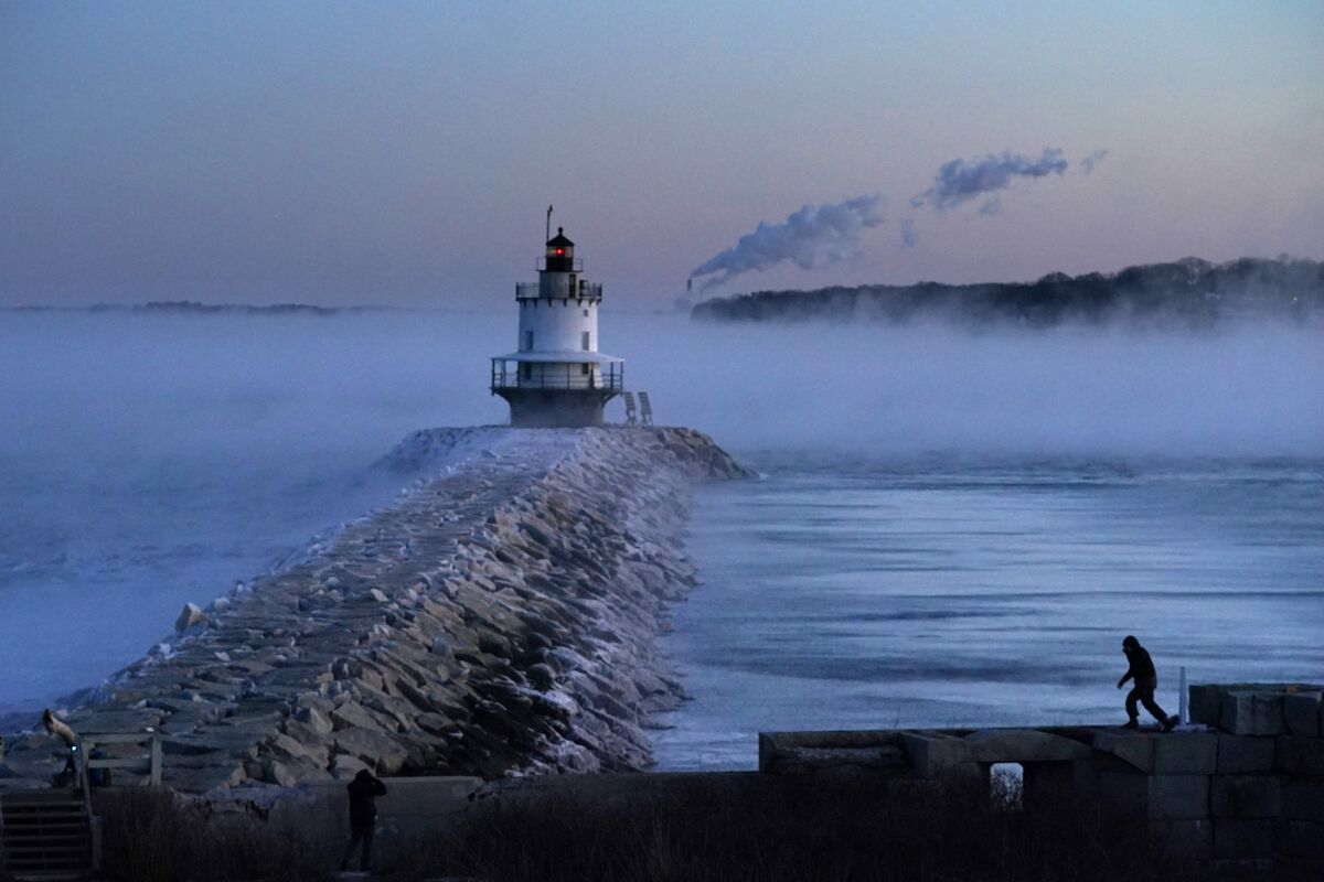 A man walks on a sea wall near a lighthouse overlooking water covered in low fog.