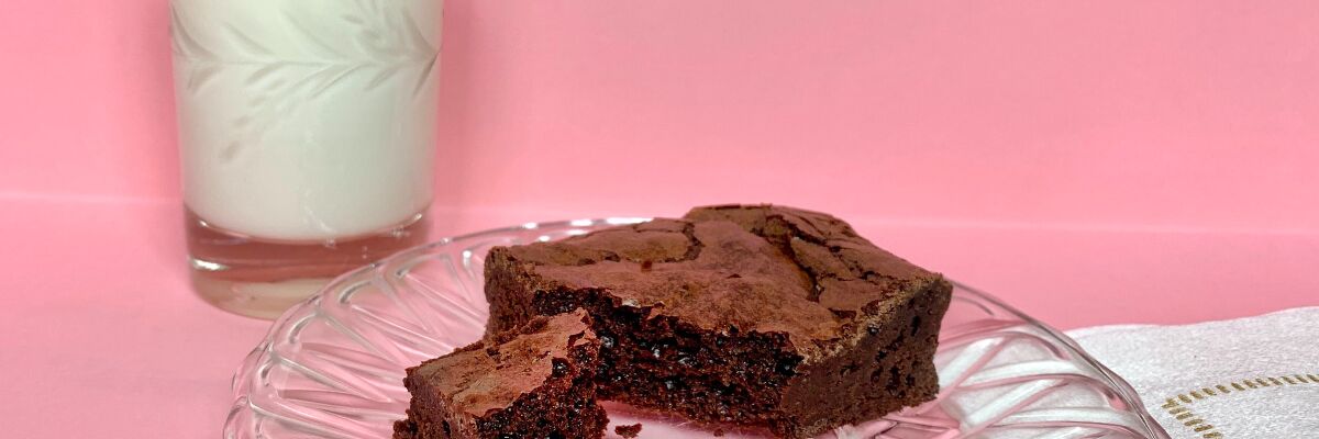 A flourless fudgy brownie with a piece broken off, on a glass plate with a glass of milk in the background