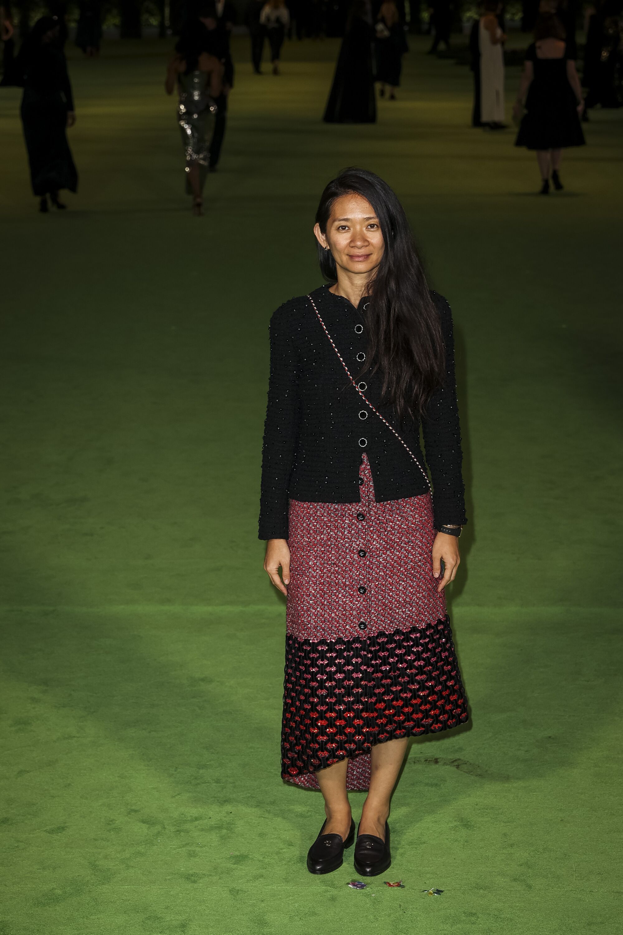 A woman in a black, button-down shirt and pink skirt posing on a green carpet