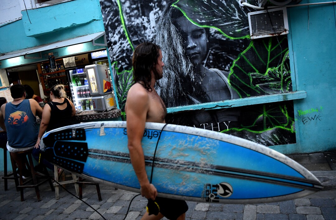 A surfer walks by a mural of Katherine Diaz.