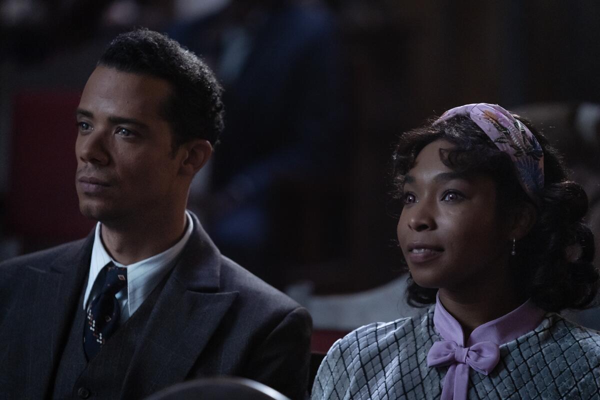 A man in a suit sits next to a little girl with a pink scarf in her hair.