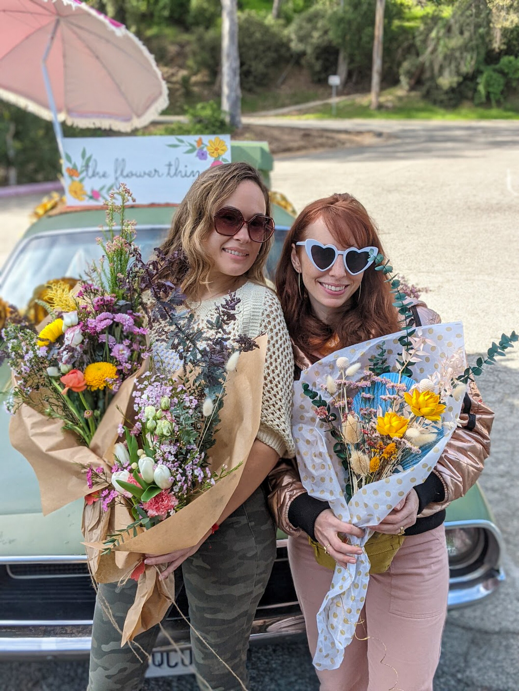 Two women lean against a car, each holding a large bouquet of flowers.