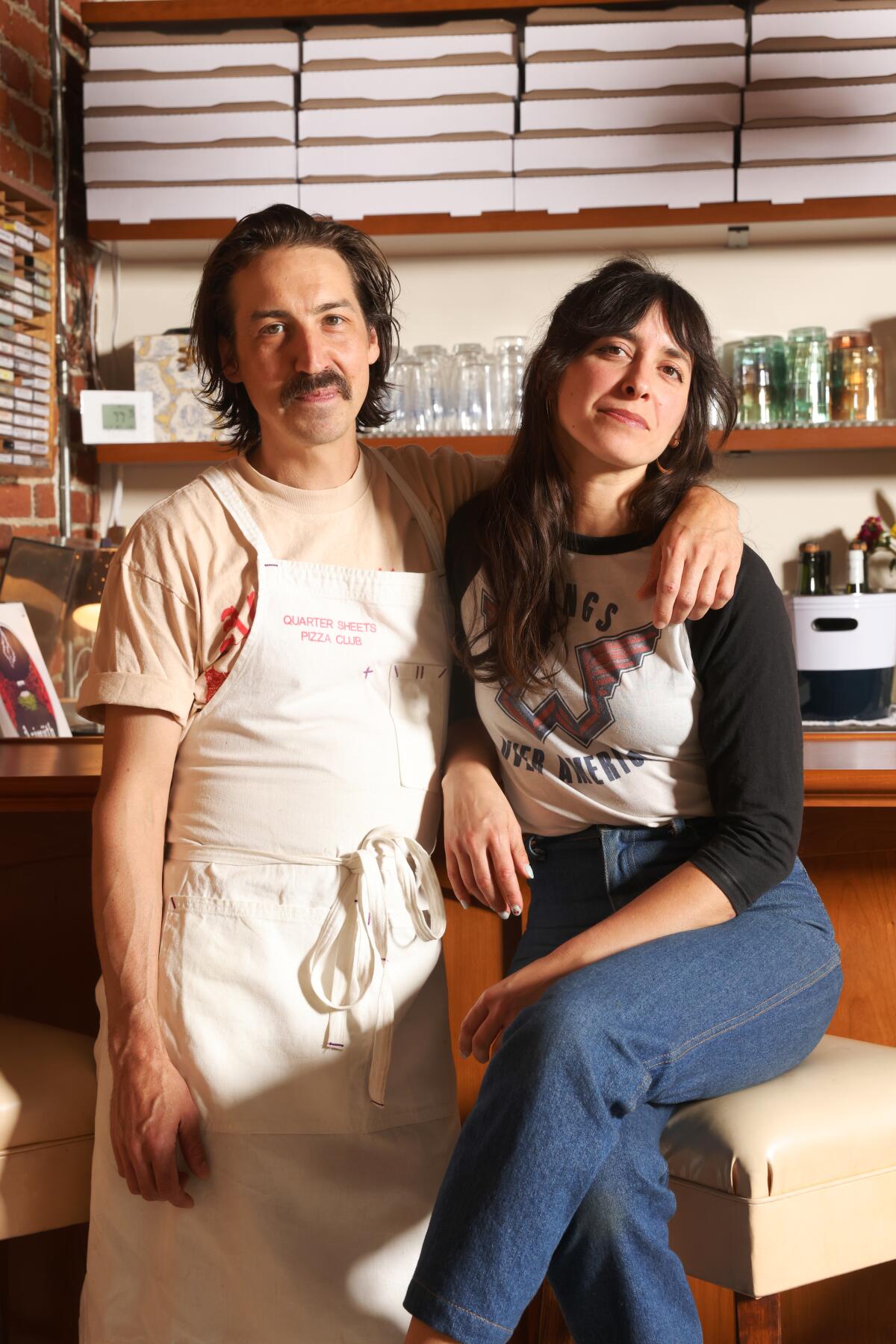 A man in a chef's apron stands next to a woman seated on a counter, his arm around her shoulder.