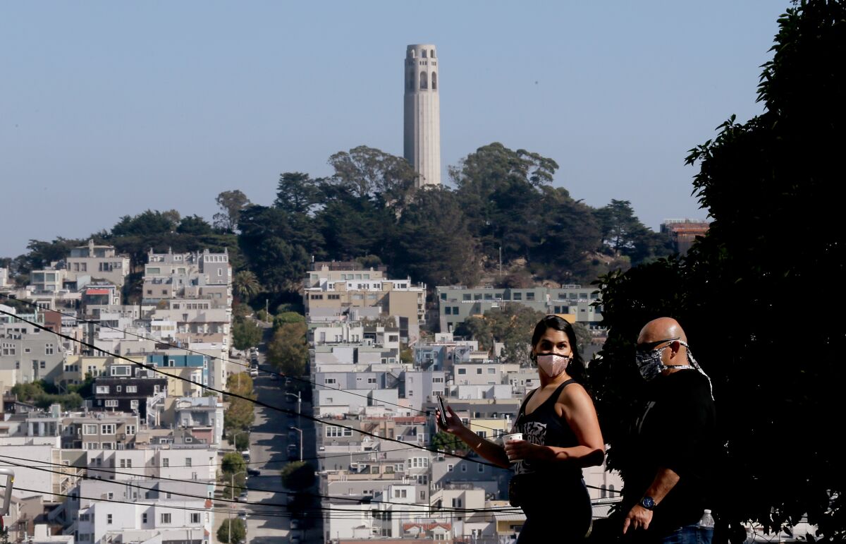Housing stacks a hillside that's topped by Coit Tower as pedestrians walk by wearing masks.