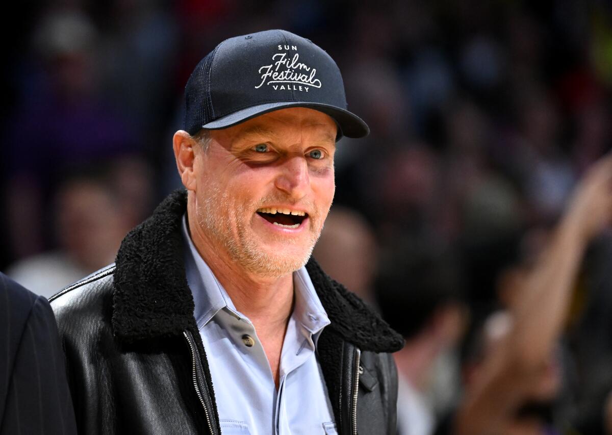 Woody Harrelson smiling at a basketball arena in a Sun Valley Film Festival hat and leather jacket