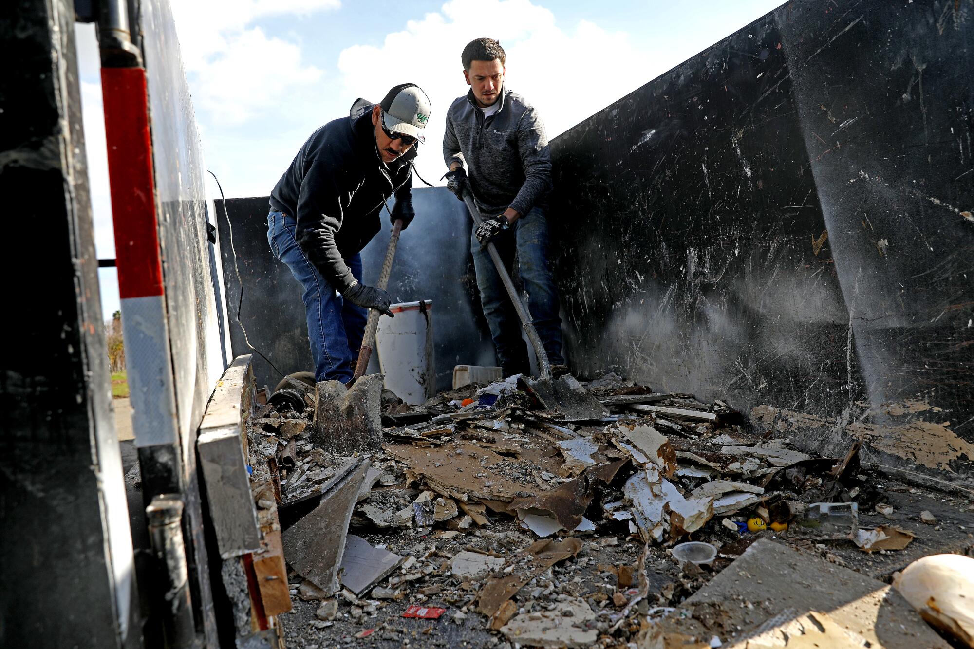 Using shovels, two men move chunks of flood debris from a trailer into a trash bin.