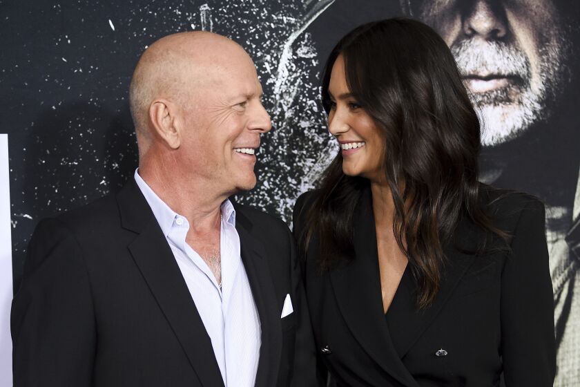 A bald man and a woman with long, brown hair smiling at each other while wearing black suits