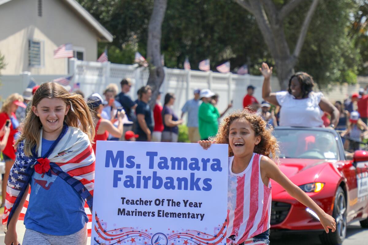 A woman stands up in a sports car behind two girls holding a banner with her name