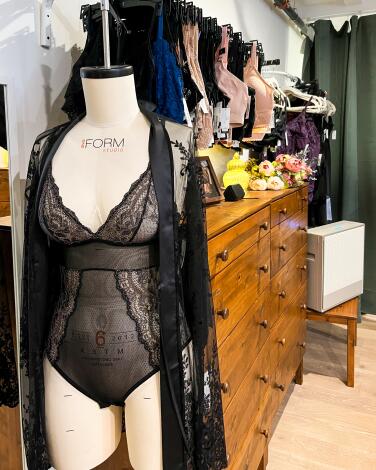 A mannequin wears a black lingerie set in front of a rack of bras. 