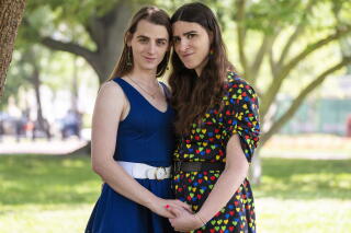 Zooey Zephyr and Erin Reed lean into each other and hold hands for a photo in a parklike setting with large trees