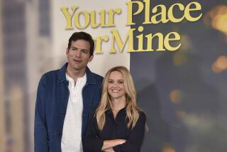 Ashton Kutcher in a blue jacket and brown pants standing next to Reese Witherspoon in a black dress
