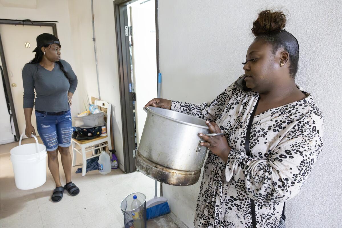 One woman holds a bucket and another holds a pot in a room.