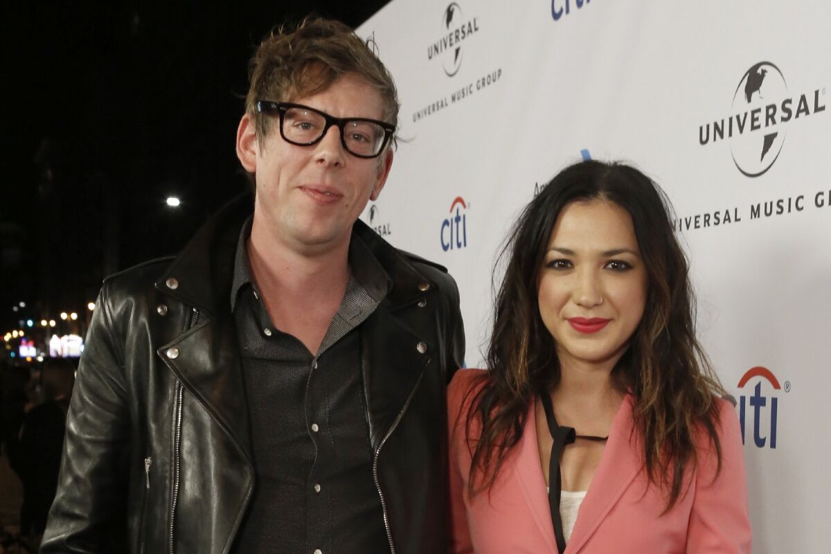 A man wearing glasses and a black leather jacket poses with a woman wearing a pink blazer