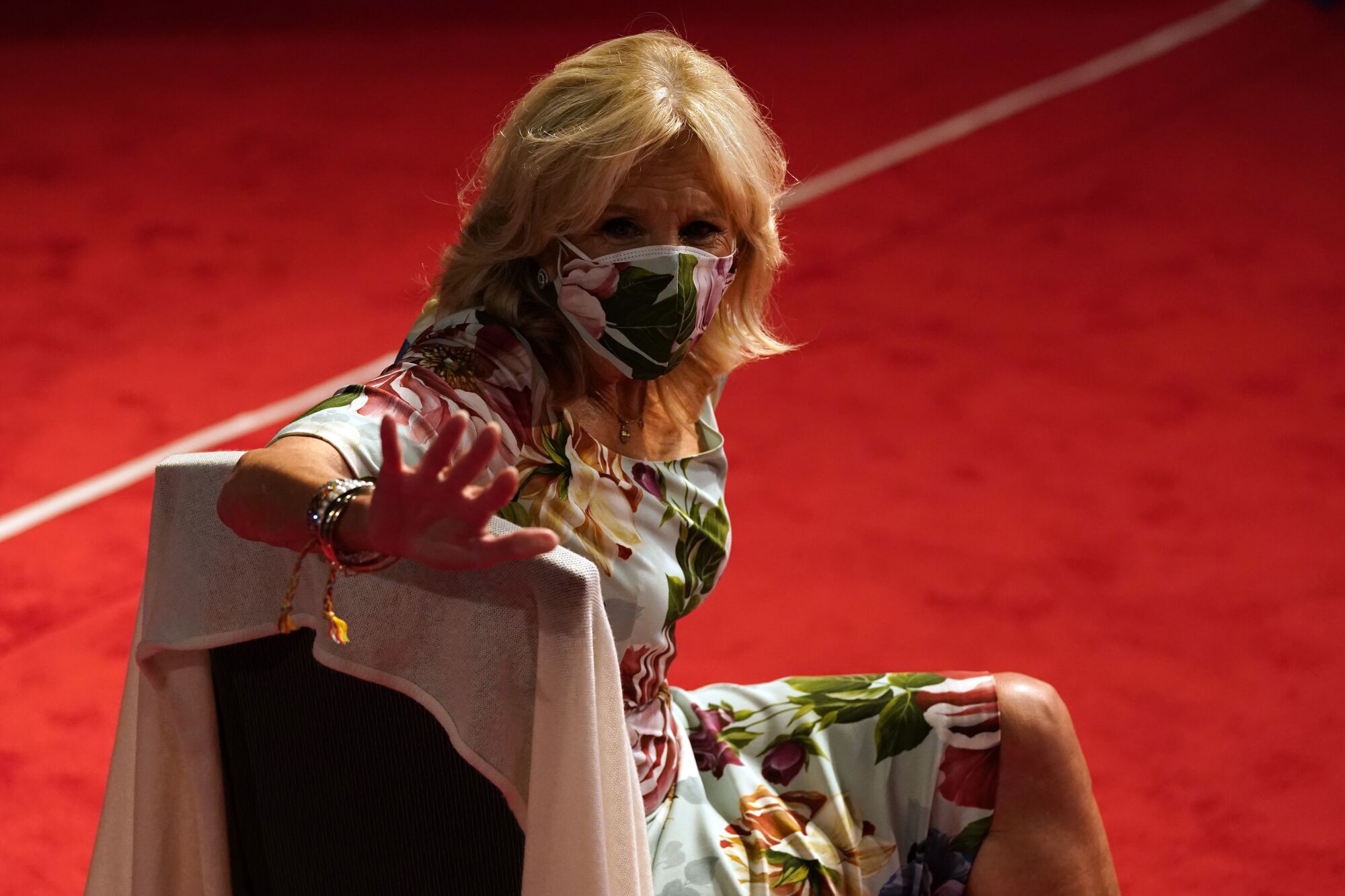 Jill Biden, wearing a floral mask that matches her dress, sits in a chair in the debate hall