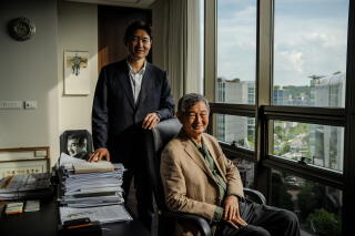 Two men pose for a portrait in an office next to a large window