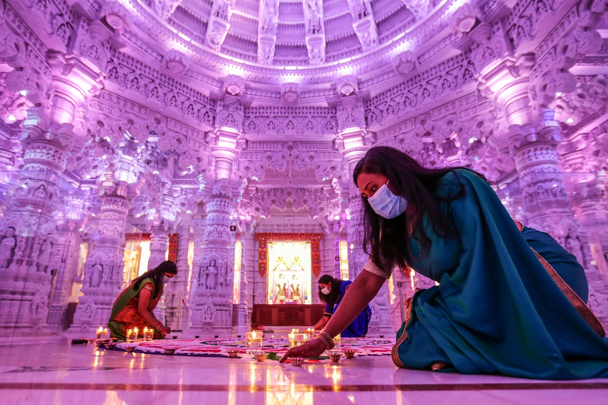 Women wearing masks and traditional robes kneel around a work of sand art inside an intricate Hindu temple