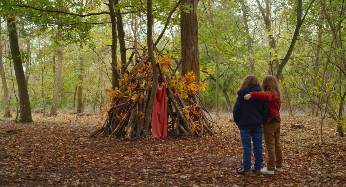 Two girls stand with their arms around each other in the woods, looking at a fort made out of sticks 