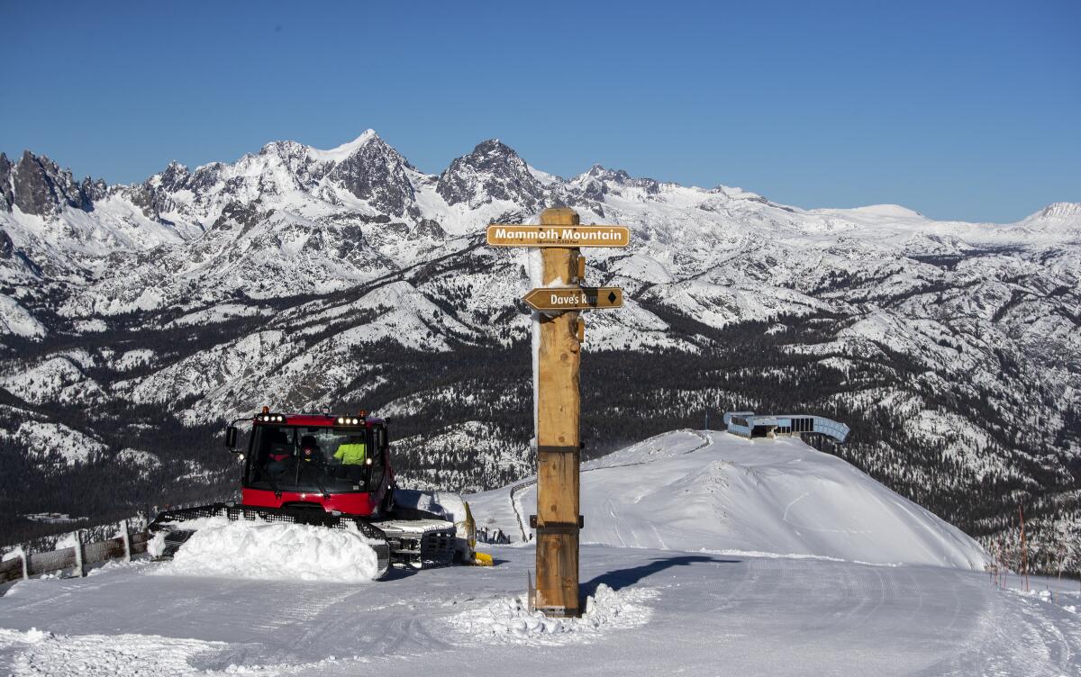A snowplow against a backdrop of jagged mountain peaks and a wooden sign that reads "Mammoth Mountain."