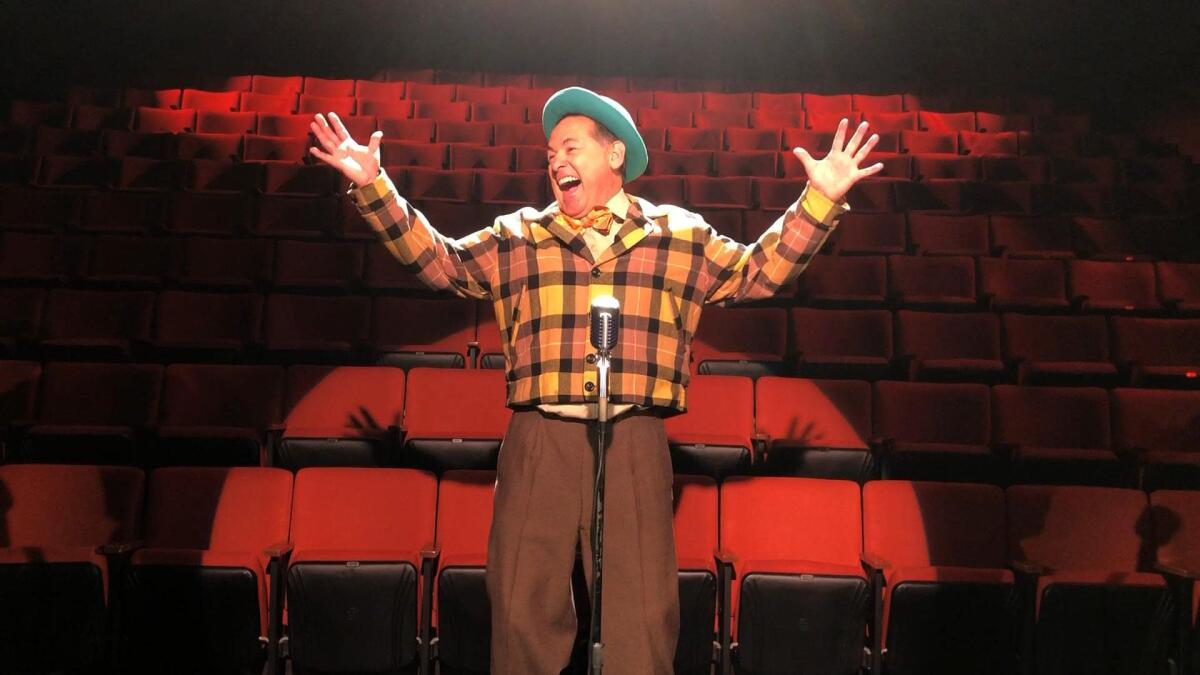 An actor stands before an empty theater, smiling and raising his hands.