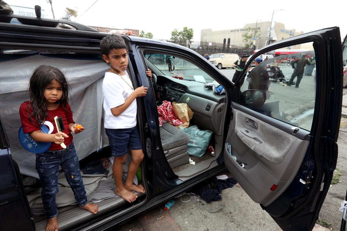 A little girl and boy stand in the open door of the car they have been sleeping in with their parents on Skid Row