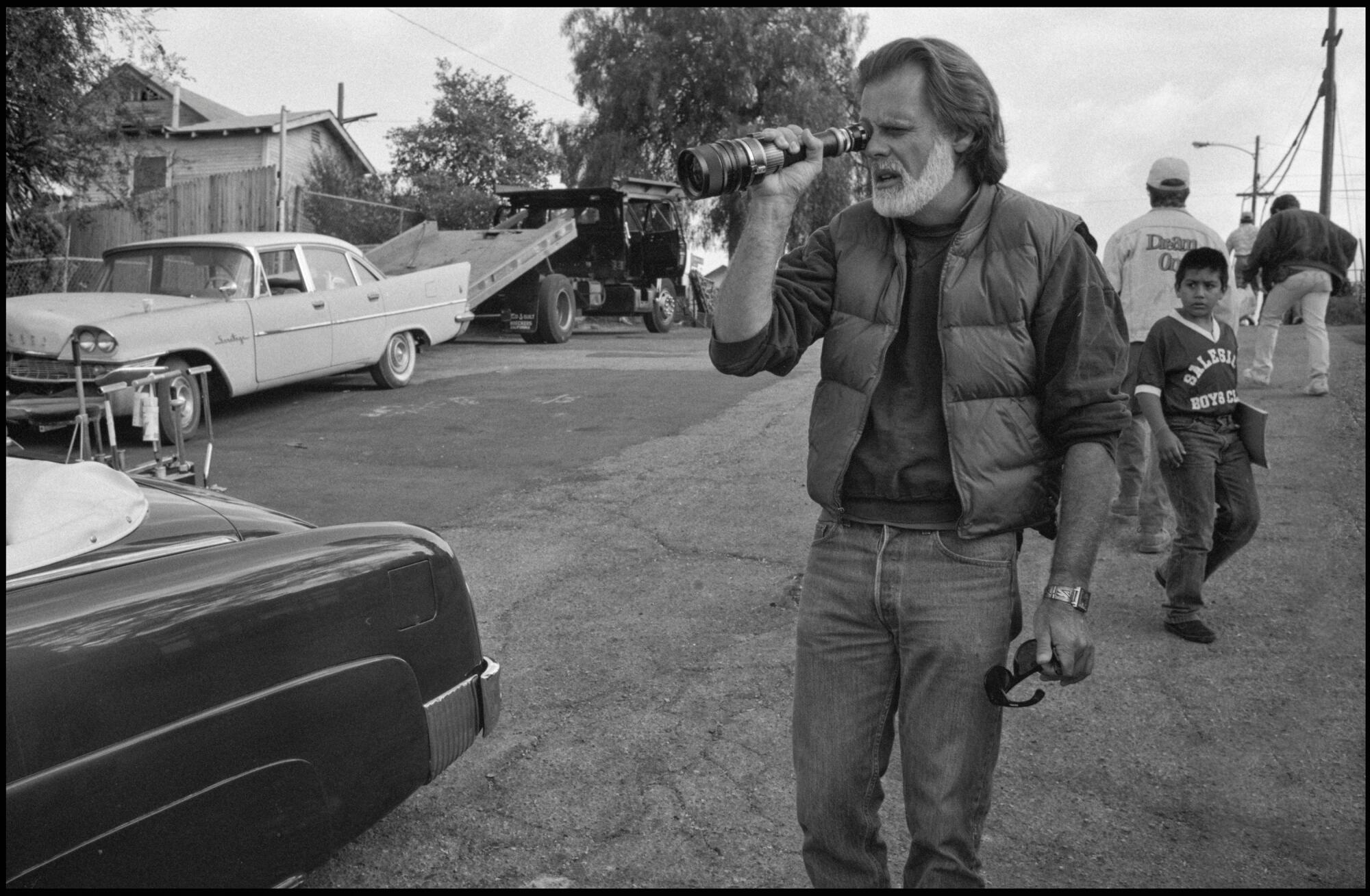 Director Taylor Hackford looks through a lens while standing next to a vintage car.