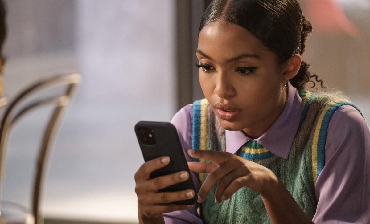 Yara Shahidi scrolls through her smartphone while wearing a colorful sweater vest.