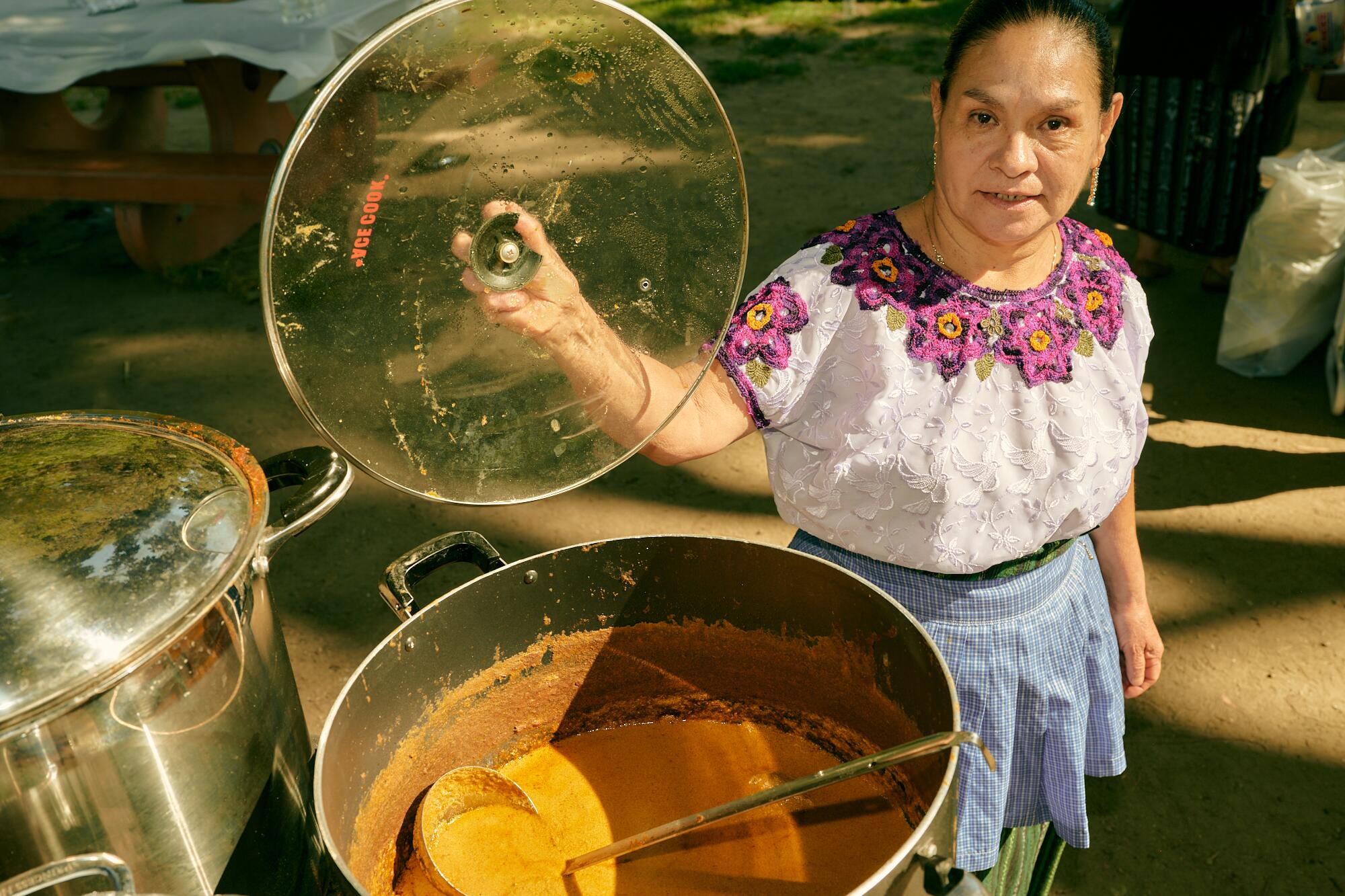 A woman lifts a glass lid to reveal a large pot of mole.