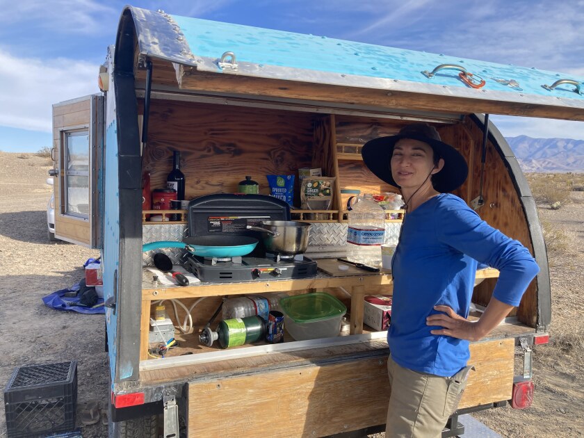 A woman in a hat and blue shirt stands before a camper whose contents include a stove and pan and kitchen supplies