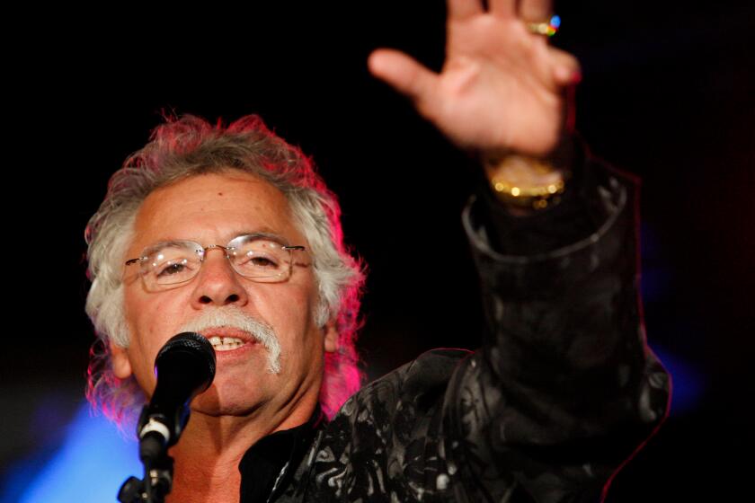 A man with greying hair and wire-framed glasses singings into a microphone with his left arm raised