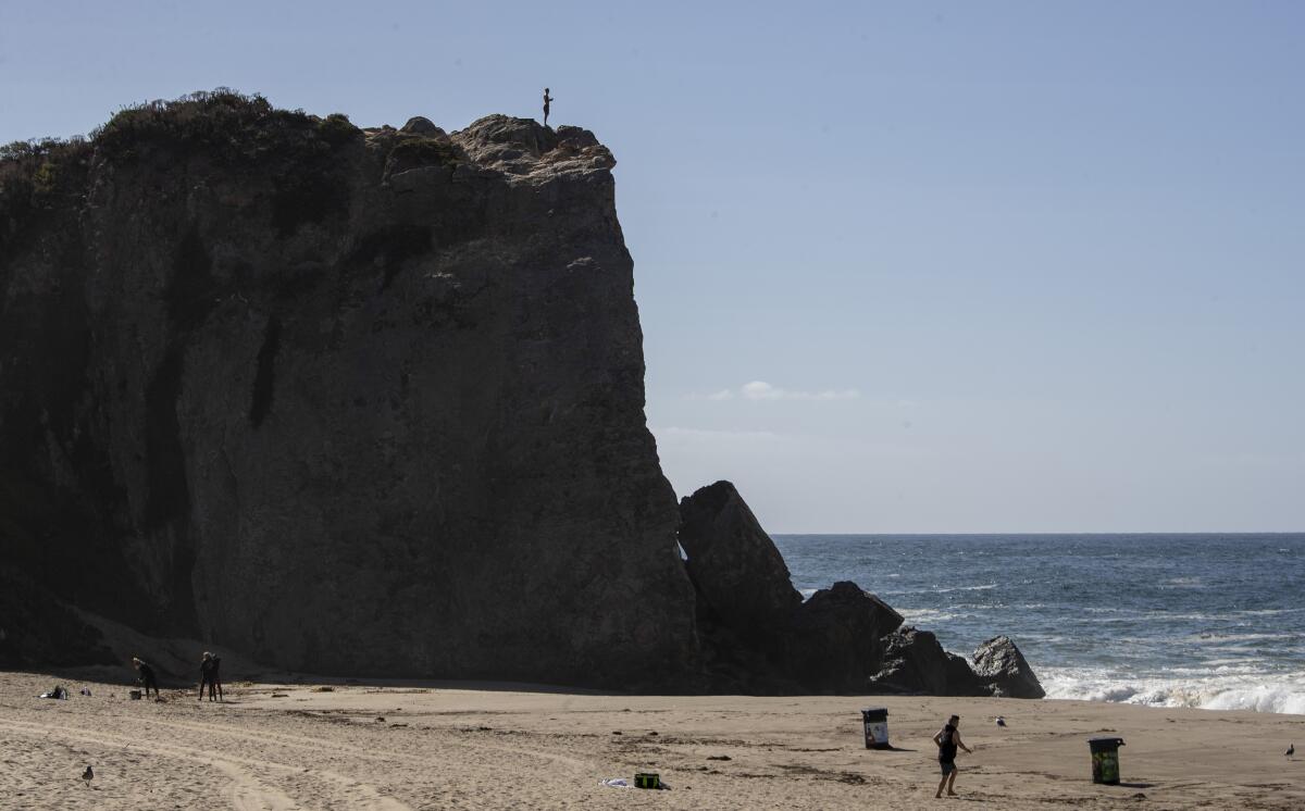 A figure stands atop a huge rock outcropping on the beach.