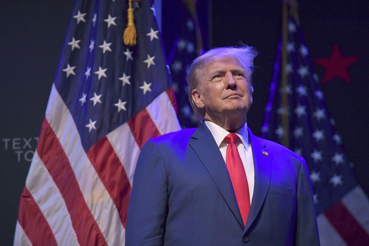 Former President Trump standing in the spotlight in front of American flags.