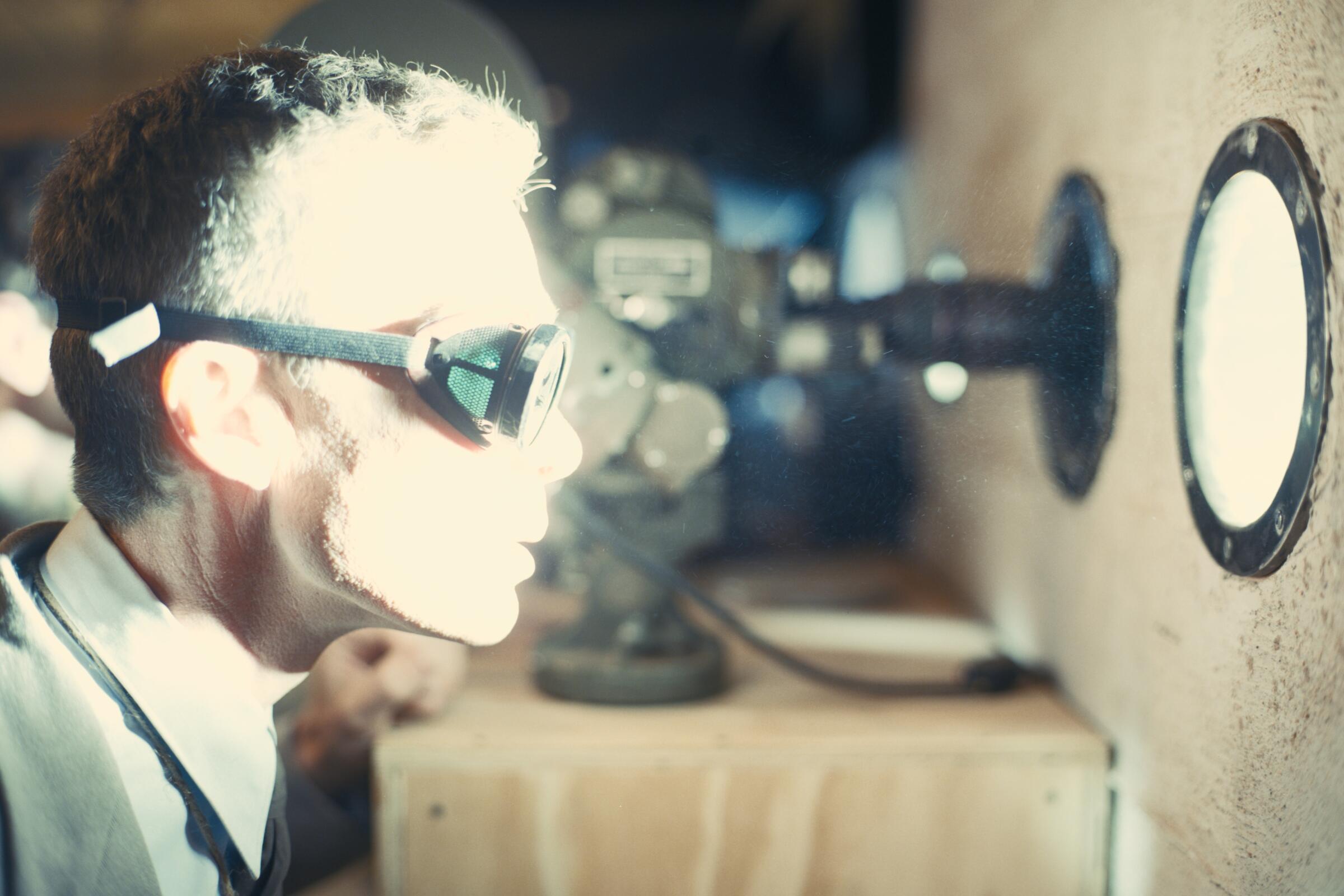A man wearing goggles looks through a small, round window as bright, white light washes over him in "Oppenheimer."