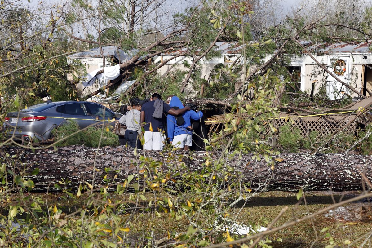 People surrounded by downed trees and branches huddle outside a tornado-damaged mobile home.