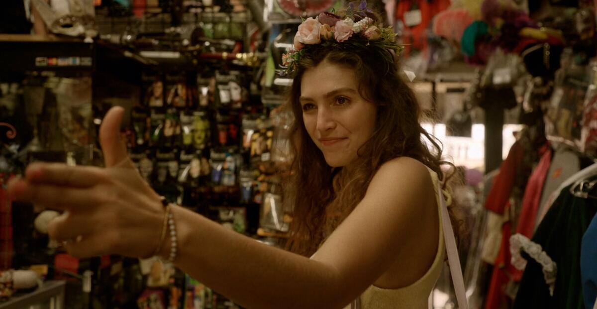A woman wearing a flower crown in a costume shop points a finger gun.