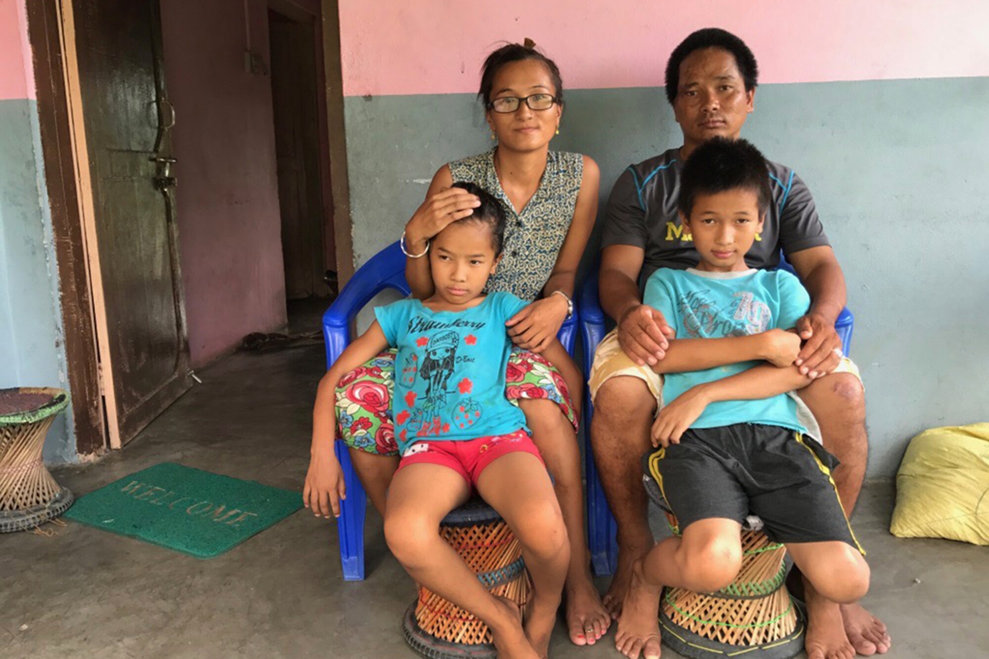 A family of four sits on plastic chairs and wicker stools on their porch