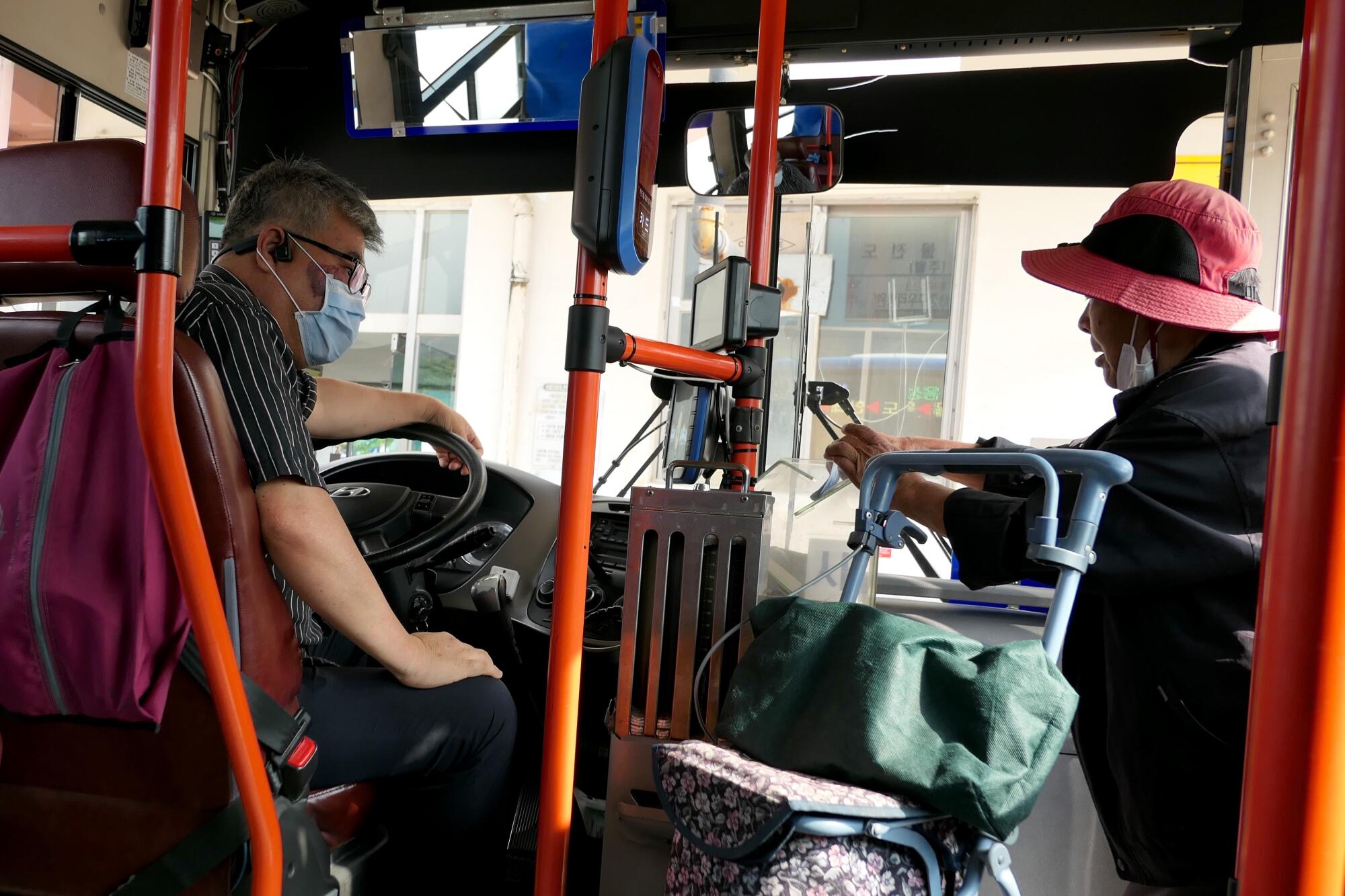 A man sits at the wheel of a bus and a woman pushing a cart gets on board. 