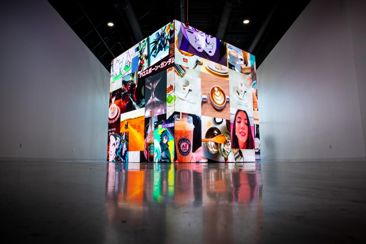 A large cube covered in video screens is filled with imagery of colorful advertisements in a white-walled space.