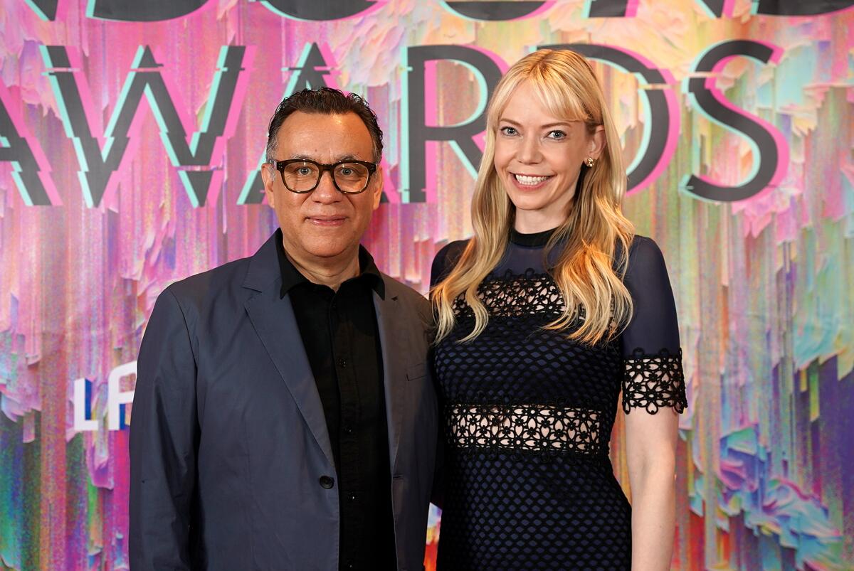 Fred Armisen, in a dark suit jacket and black shirt, smiles standing with Riki Lindhome, clad in a black lace dress 