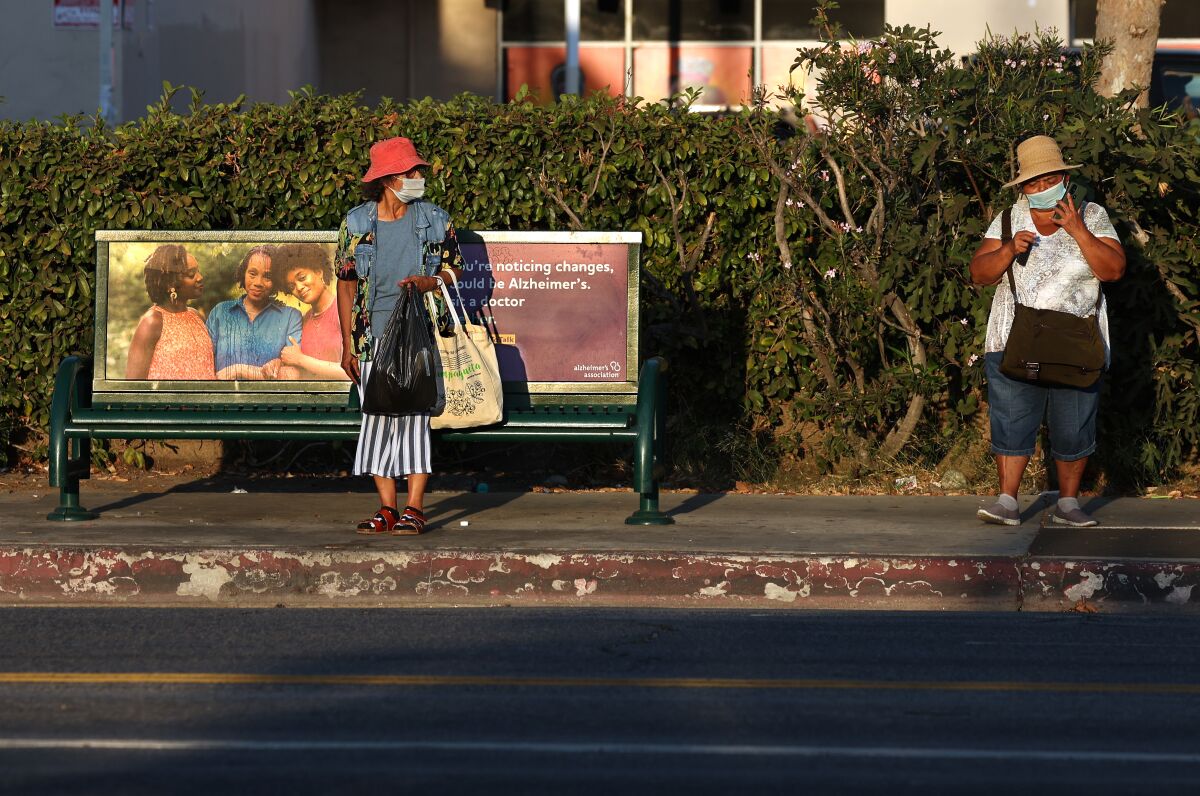 Two women wearing face masks stand at a bus stop in Van Nuys in the afternoon sun.