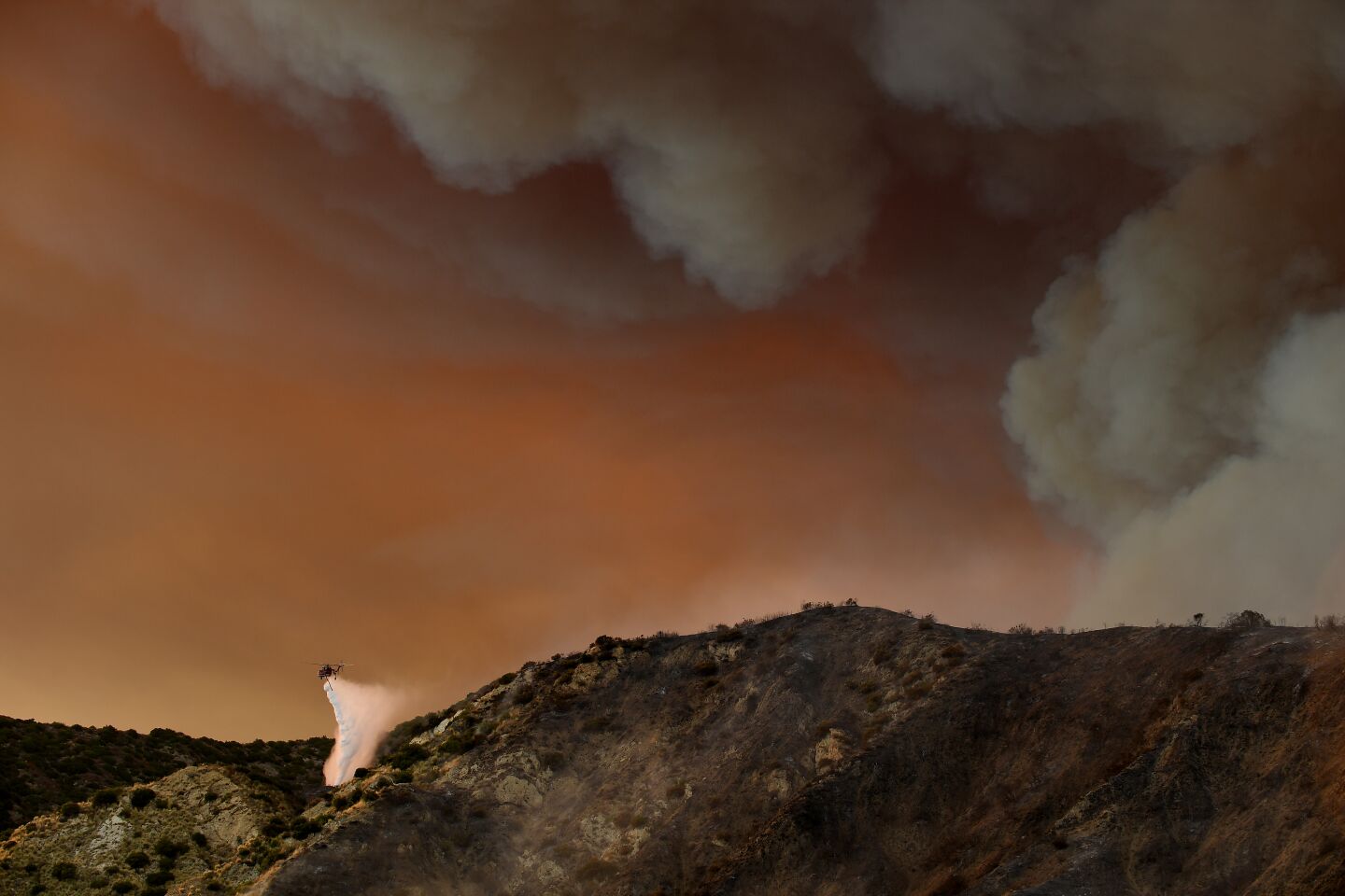 Smoke fills the sky as a helicopter makes a water drop on the Ranch fire in the hills of Azusa