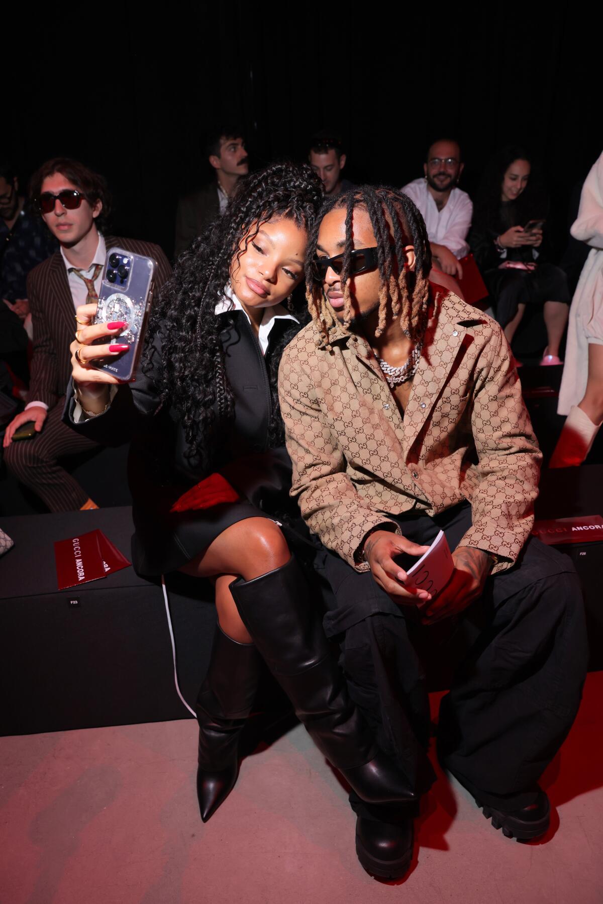 Halle Bailey in a black outfit and boots posing for a seated selfie with a man with braids, sunglasses and a brown jacket 