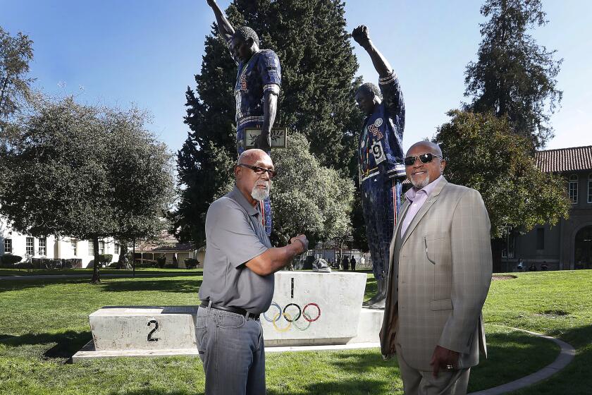 FILE - In this Oct. 17, 2018, file photo, John Carlos, left, and Tommie Smith pose for a photo in front of statue that honors their iconic, black-gloved protest at the 1968 Olympic Games, on the campus of San Jose State University in San Jose, Calif. The U.S. Olympic and Paralympic Committee heeded calls from American athletes, announcing Thursday, Dec. 10, 2020, that it won’t sanction them for raising their fists or kneeling on the medals stand at next year’s Tokyo Games and beyond. (AP Photo/Tony Avelar, File)