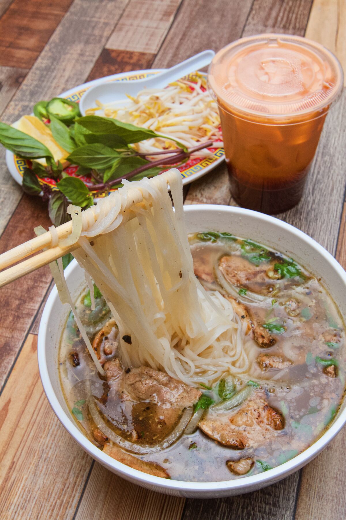 Chopsticks lift rice noodles from charred-pork pho. Behind is a cup of Thai iced tea and a plate of basil and bean sprouts.