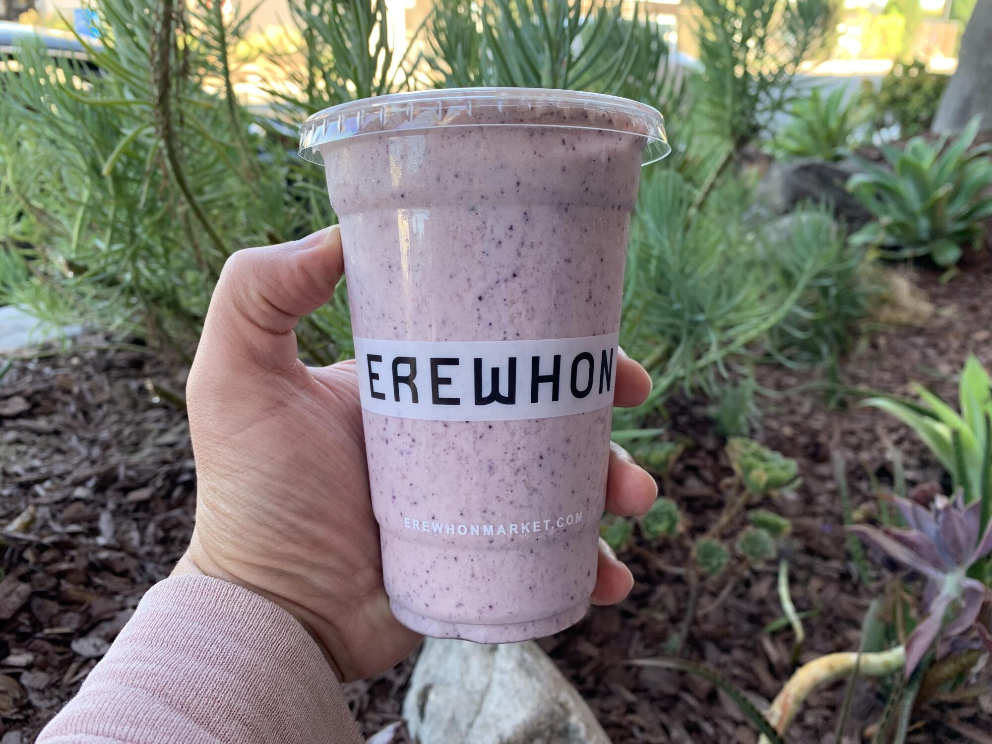 A hand holds a purple smoothie in a plastic cup that says Erewhon.