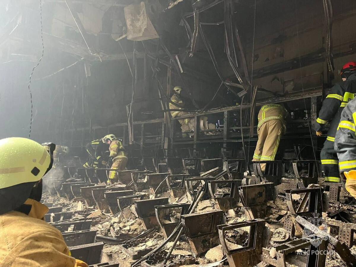 Emergency workers wearing yellow coats and helmets walk among burned seats in a concert hall. 